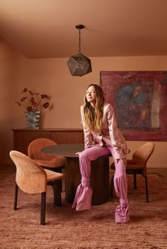 a woman sitting on top of a wooden table in front of a painting and chairs