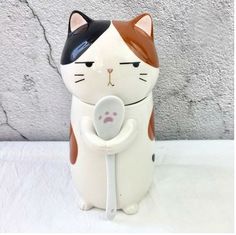 a ceramic cat with a spoon in its mouth sitting next to a wall and cement background