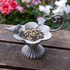 a bird feeder sitting on top of a wooden table with flowers in the back ground