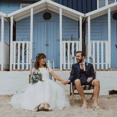 a bride and groom sitting on a chair in front of blue beach huts holding hands