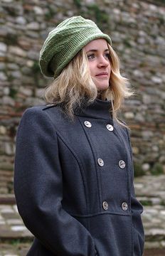 a woman standing in front of a stone wall wearing a green hat and black coat