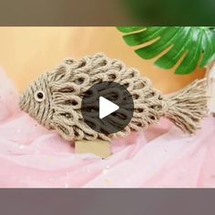 a fish made out of rope sitting on top of a pink cloth next to a green plant