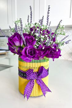 purple flowers are in a yellow container on a counter top, tied with a purple ribbon