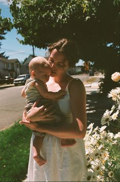 a woman holding a baby in her arms on the side of a road next to flowers