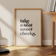 a framed poster with the words take a seat, sweet cheeks on it next to a wooden table