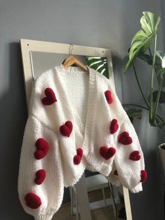 a white sweater with red hearts on it hanging from a mirror next to a potted plant