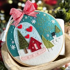 an embroidered christmas ornament with snowmen and trees on it, sitting on a wooden tray