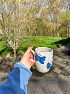 a woman holding a coffee cup with blue butterflies painted on it in front of some trees