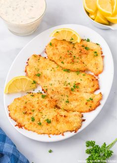 three fried fish fillets on a white plate with lemon wedges and parsley