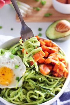 someone is holding a fork over a bowl of green spaghetti with carrots and avocado