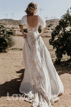 the back of a woman in a white wedding dress standing on a dirt road with her hands behind her back