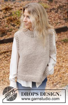 a woman wearing a sweater and jeans standing in front of some trees with leaves on the ground