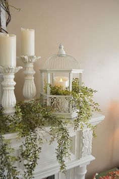 a white birdcage with greenery and two candles on top of the mantle