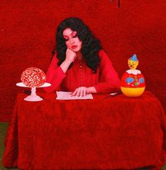 a woman sitting at a table with two cakes on top of it and a red wall behind her