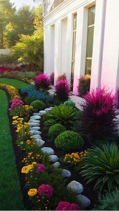 a garden with flowers and rocks in the grass next to a white house on a sunny day