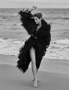 a woman is walking on the beach with her arms in the air while wearing an ostrich feather coat