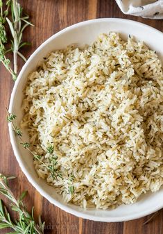 a white bowl filled with rice on top of a wooden table next to some herbs