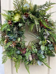 a wreath with green plants and other greenery hanging on the front door to welcome guests