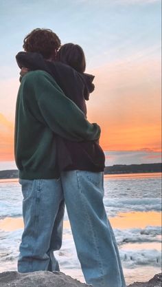 two people standing next to each other on top of a rock near the ocean at sunset