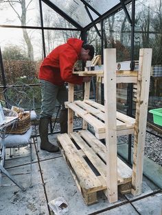 a man in red jacket and boots working on a wooden shelf inside of a greenhouse