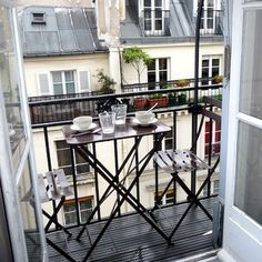 a balcony with two small tables and chairs on it, overlooking the city from an open window