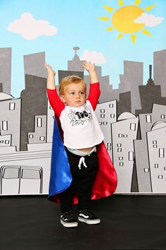 a little boy dressed as superman standing in front of a cityscape backdrop with his hands up