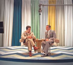 two men sitting on chairs in front of colorful drapes, talking to each other