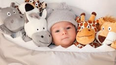 a baby laying in bed with stuffed animals on it's back and wearing a hat