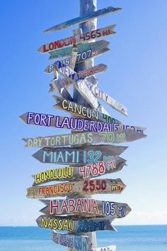 a sign post with many different signs on it in front of the ocean and blue sky