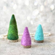 three small felted trees sitting on top of a white table next to a beaded necklace