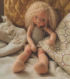 a doll sitting on top of a bed next to a pillow and throw blanket in front of it