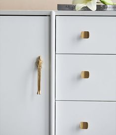 a white dresser with two drawers and a gold handle on the top drawer, next to a flower