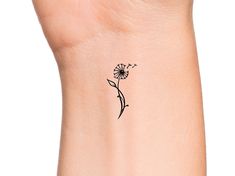 a small black flower tattoo on the left side of the ankle, with a single dandelion coming out of it