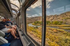 two people sitting on a train looking out the window at a river and mountains in the distance