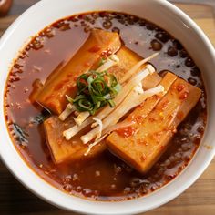 a bowl filled with tofu and sauce on top of a wooden table next to a spoon