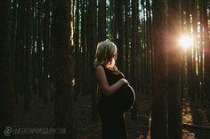 a pregnant woman standing in the middle of a forest at sunset with the sun shining through the trees