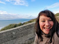 a woman smiling while standing next to a wooden fence near the ocean on a sunny day