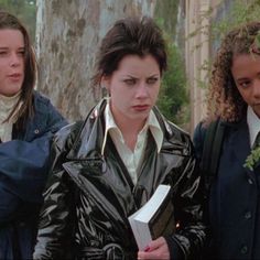 three young women standing next to each other in front of a tree and one holding a book