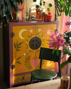 a green chair sitting in front of a yellow cabinet with flowers on it and a potted plant next to it