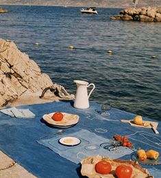 an outdoor table with plates and bowls on it near the water, next to some rocks