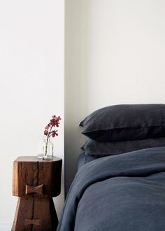 a bed with blue sheets and pillows next to a small wooden table holding a flower