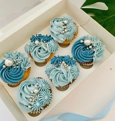 four cupcakes with blue frosting and flowers in a box on a table