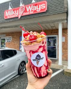 a person holding up a milkshake in front of a building with cars parked outside