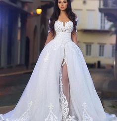 a woman wearing a white wedding gown with sheer sleeves and high slited skirt is standing in front of a building