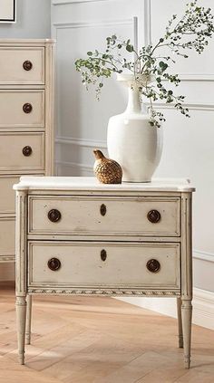 a white dresser with drawers and a vase on it's top, sitting in a room