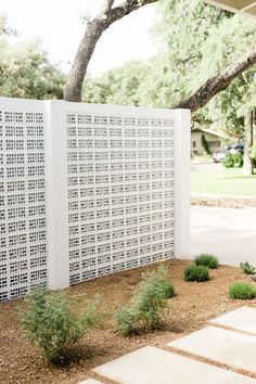 a white privacy fence in front of a tree and shrubbery on the side of a house