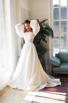 a woman in a white wedding dress is standing by a window with her arms behind her head