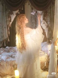 a woman with long hair standing in front of a bed wearing a white dress and holding her hand up to the ceiling