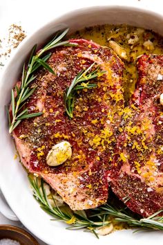 two pieces of meat in a white bowl with herbs and seasoning on top, surrounded by salt and pepper