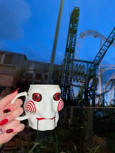 a hand holding a coffee mug with a face painted on it in front of a roller coaster